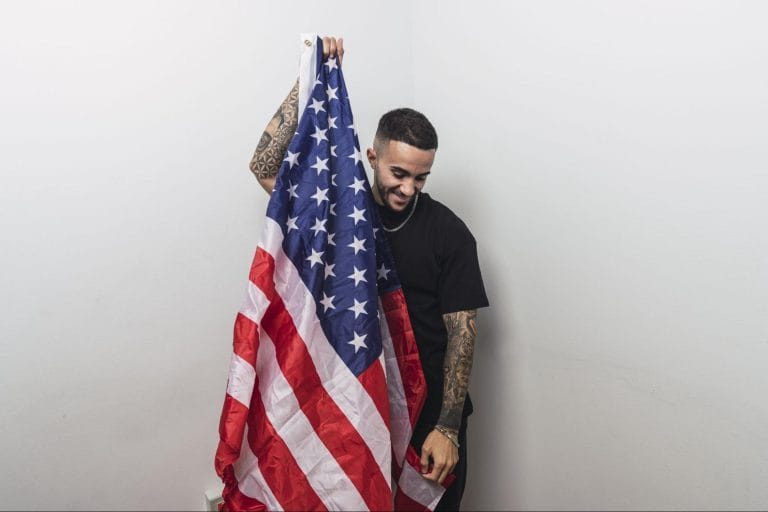 A smiling man with tattoos holding a large American flag against a plain white wall.