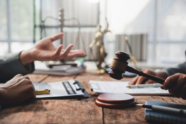A wooden gavel being held over a desk during a legal discussion with contracts and a Lady Justice statue.
