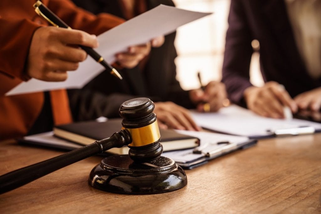 A close-up of a wooden gavel and sound block on a desk in the foreground, with the blurred figures of legal professionals in the back.